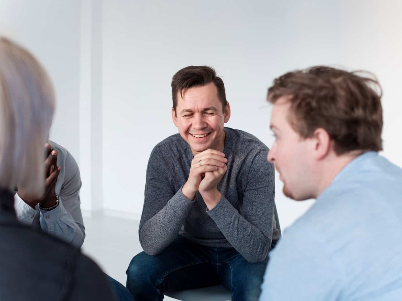 Ein Symbolbild für eine Selbsthilfegruppe oder Therapie. Ein Mann mit kurzen dunklen Haaren sitzt in einem Stuhlkreis und blickt lächelnd und aufmerksam zu einer anderen Person. Die übrigen Teilnehmer der Gruppe sind im Vordergrund nur unscharf von hinten oder von der Seite zu sehen.
