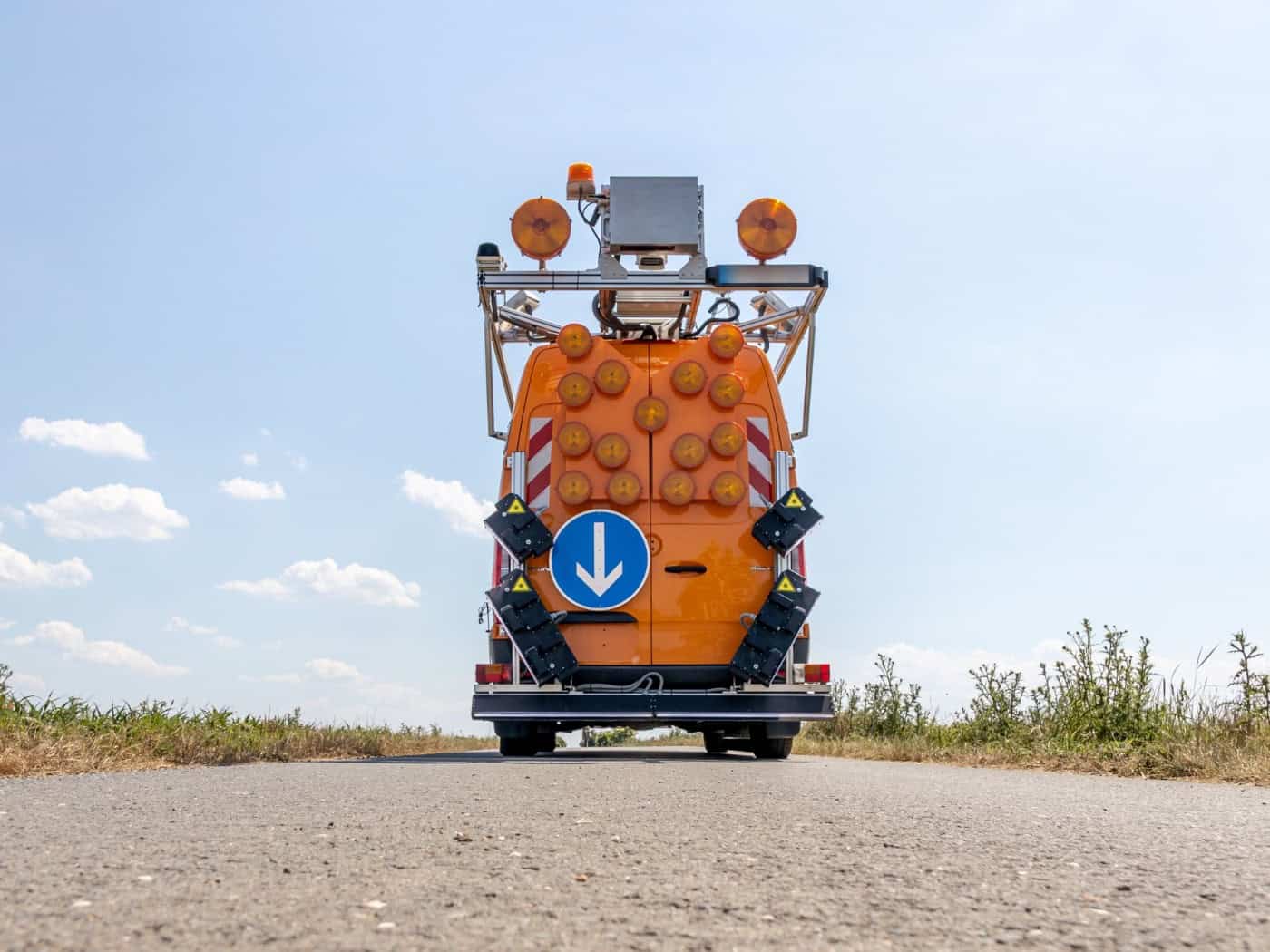 Die Heckansicht eines orangefarbenen Messbusses, ausgestattet mit zahlreichen runden Warnleuchten, einem blauen Pfeilschild nach unten und komplexen Laserscannern an den Seiten, aufgenommen aus einer tiefen Perspektive auf dem Asphalt.