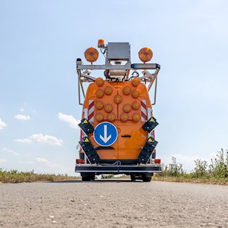 Die Heckansicht eines orangefarbenen Messbusses, ausgestattet mit zahlreichen runden Warnleuchten, einem blauen Pfeilschild nach unten und komplexen Laserscannern an den Seiten, aufgenommen aus einer tiefen Perspektive auf dem Asphalt.