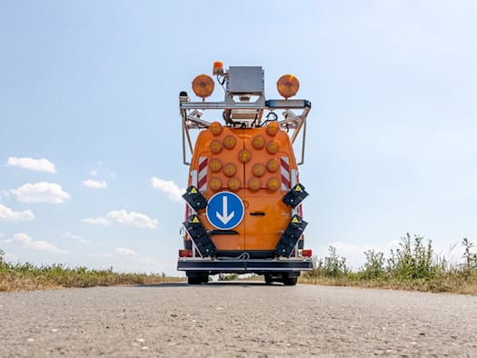 Die Heckansicht eines orangefarbenen Messbusses, ausgestattet mit zahlreichen runden Warnleuchten, einem blauen Pfeilschild nach unten und komplexen Laserscannern an den Seiten, aufgenommen aus einer tiefen Perspektive auf dem Asphalt.