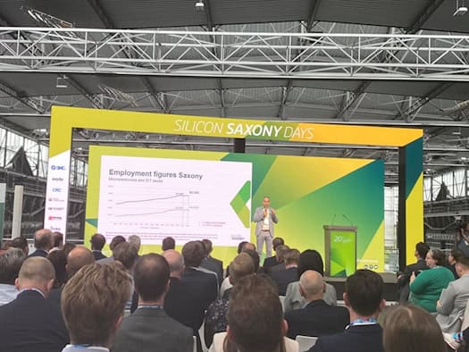 Foto aus der Perspektive des Publikums in einer modernen Eventhalle mit einer industriellen Stahlträger-Dachkonstruktion. Auf der Bühne steht ein Redner neben einer großen Präsentationswand, die die Grafik „Employment figures Saxony“ und statistische Daten zum Beschäftigungswachstum zeigt.