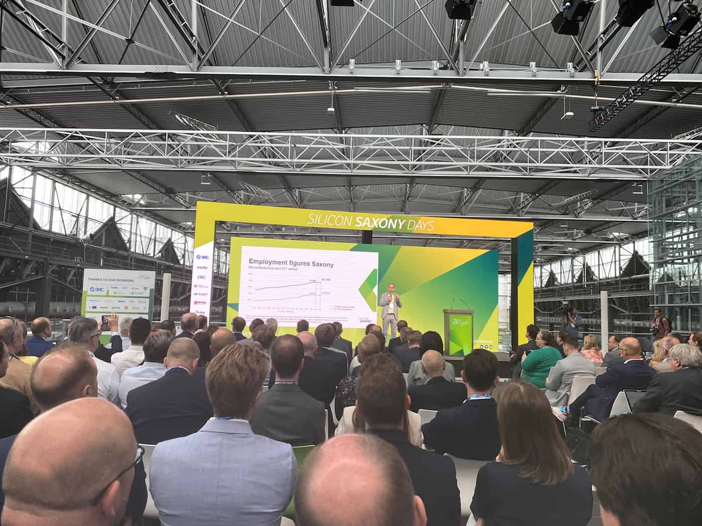 Foto aus der Perspektive des Publikums in einer großen Eventhalle mit einer industriellen Stahlträger-Dachkonstruktion. Auf der Bühne steht neben der Aufschrift „SILICON SAXONY DAYS“ ein Redner im hellen Anzug neben einer großen Präsentationswand, die eine Grafik mit der Überschrift „Employment figures Saxony“ und statistische Daten zum Beschäftigungswachstum zeigt. 