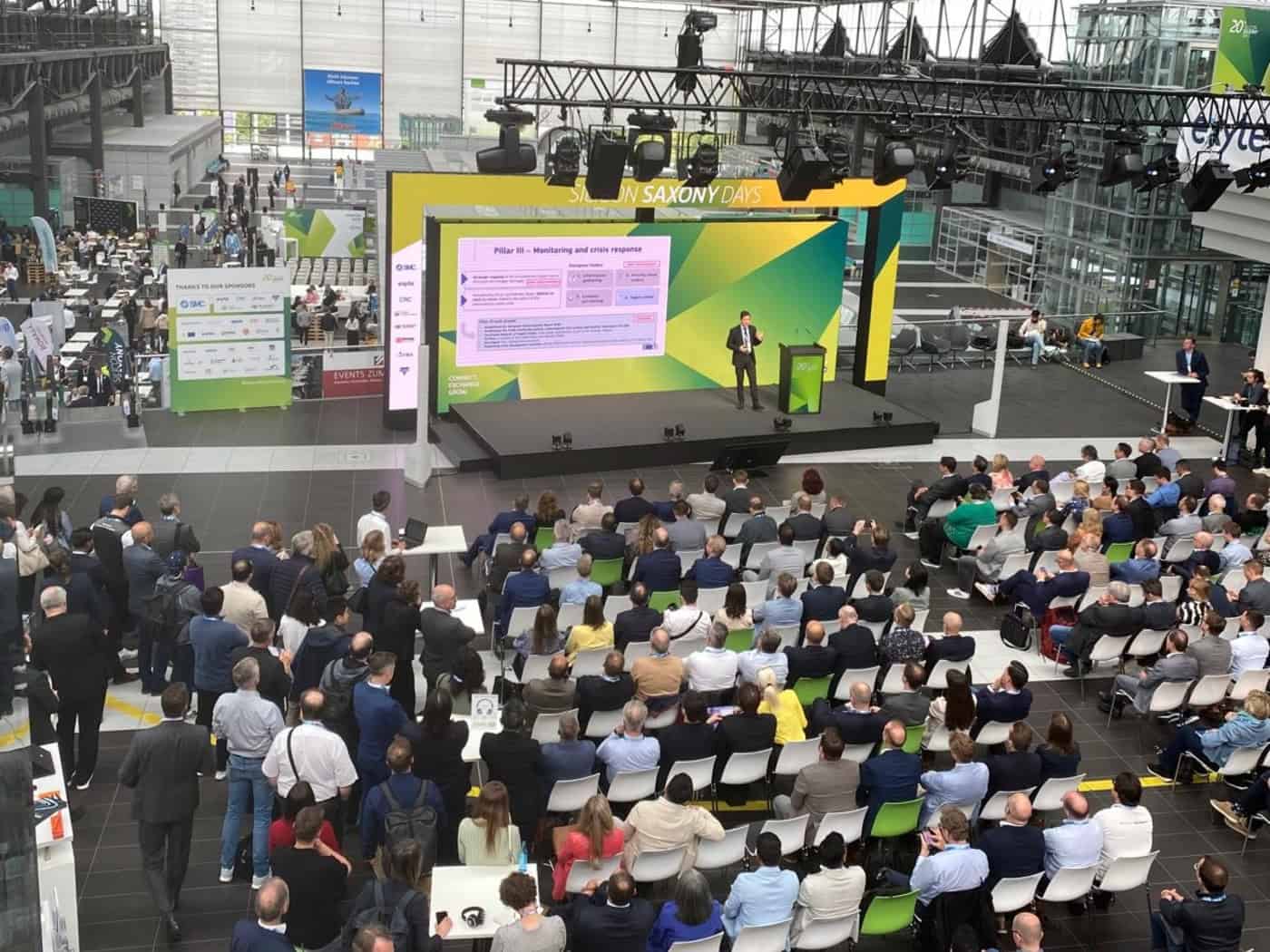 Blick von oben auf eine große Eventhalle mit Stuhlreihen voller Publikum. Auf der grün-gelben Bühne steht ein Redner vor einer Präsentation mit dem Titel „Pillar III“. Links im Hintergrund sind Ausstellungsstände erkennbar.