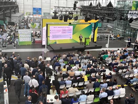 Blick von oben auf eine große Eventhalle mit Stuhlreihen voller Publikum. Auf der grün-gelben Bühne steht ein Redner vor einer Präsentation mit dem Titel „Pillar III“. Links im Hintergrund sind Ausstellungsstände erkennbar.