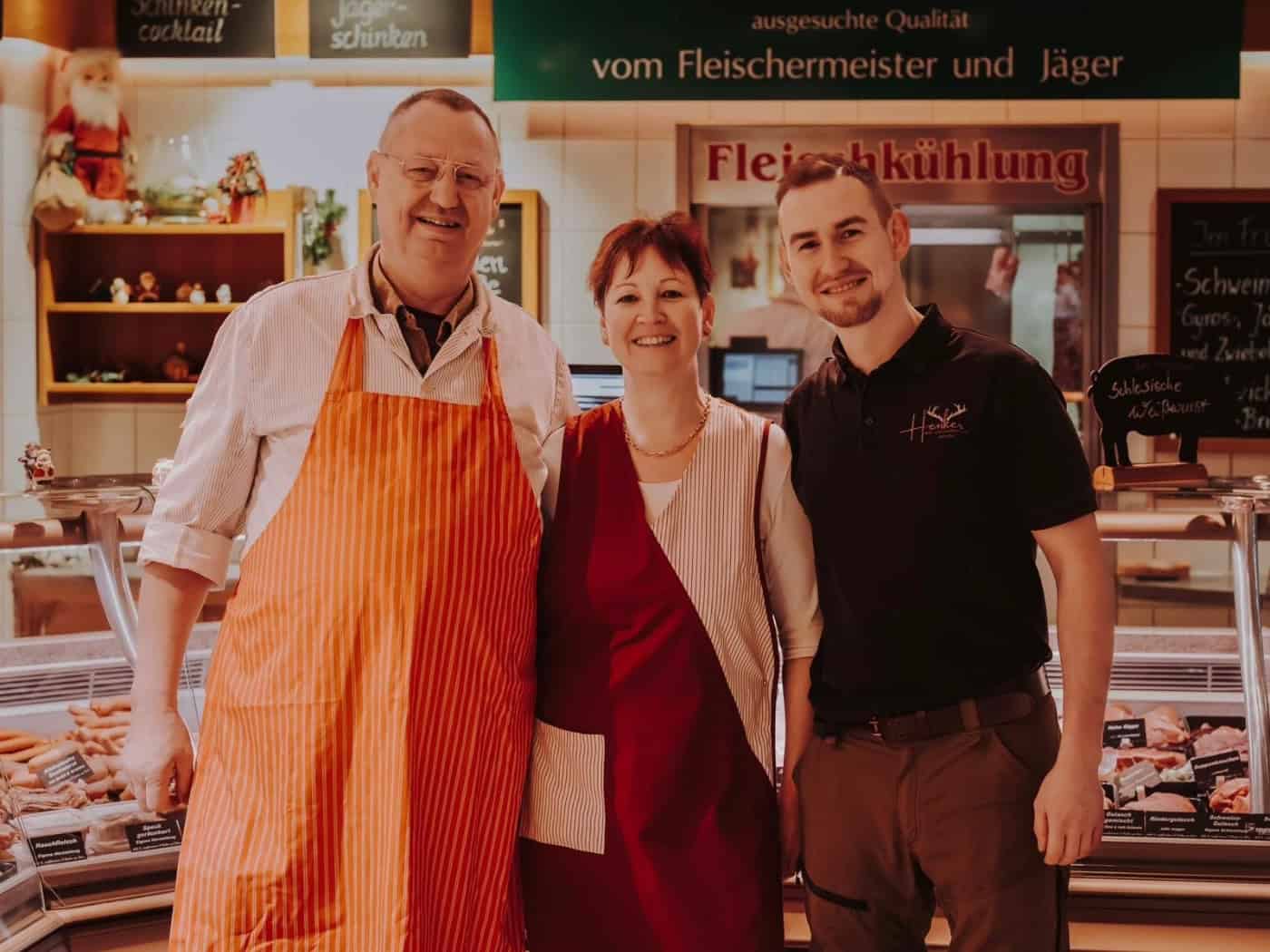 Foto: Ein älterer Mann, eine Frau in Schürzen und ein jüngerer Mann mit Hose und T-Shirt stehen lächelnd in die Kamera vor einer Theke einer Fleischerei.