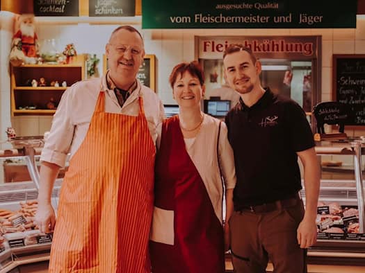 Foto: Ein älterer Mann, eine Frau in Schürzen und ein jüngerer Mann mit Hose und T-Shirt stehen lächelnd in die Kamera vor einer Theke einer Fleischerei.