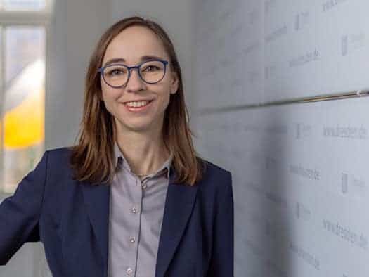 Porträt von Dr. Kristin Klaudia Kaufmann, der Sozialbürgermeisterin von Dresden, in einem blauen Sakko und mit Brille vor einer Wand mit dem Logo der Landeshauptstadt.