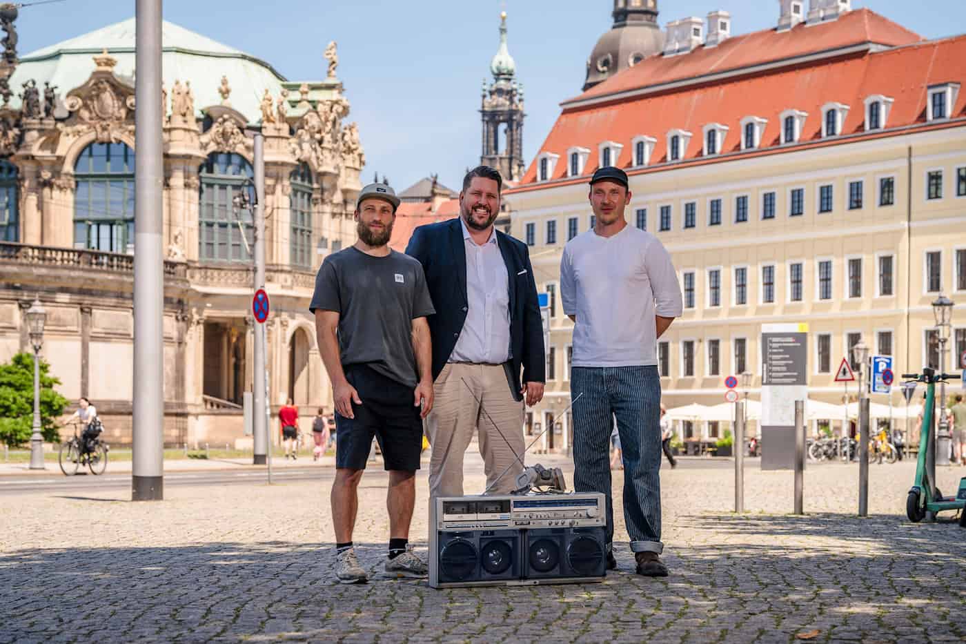 Felix Roßberg, Henrik Oliver von Oehsen und Enrico Sutter stehen lächelnd auf einem gepflasterten Weg in Dresden. Im Vordergrund auf dem Boden steht ein großer, silberner Retro-Ghettoblaster. Im Hintergrund ist die barocke Fassade des Dresdner Zwingers zu sehen.