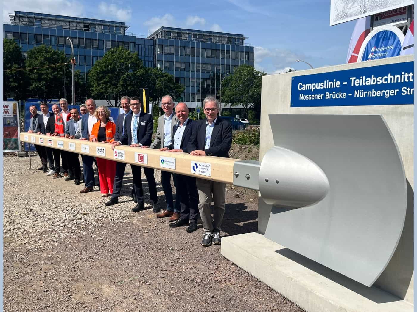 Eine Gruppe von Vertretern aus Politik und Wirtschaft steht in einer Reihe auf Schotterboden und hält gemeinsam den extrem langen Holzstiel einer überdimensionalen, silbernen Schaufel. Rechts im Bild ist eine Betonwand mit der blauen Aufschrift „Campuslinie - Teilabschnitt Nossener Brücke - Nürnberg