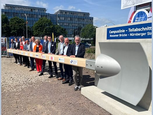 Eine Gruppe von Vertretern aus Politik und Wirtschaft steht in einer Reihe auf Schotterboden und hält gemeinsam den extrem langen Holzstiel einer überdimensionalen, silbernen Schaufel. Rechts im Bild ist eine Betonwand mit der blauen Aufschrift „Campuslinie - Teilabschnitt Nossener Brücke - Nürnberg