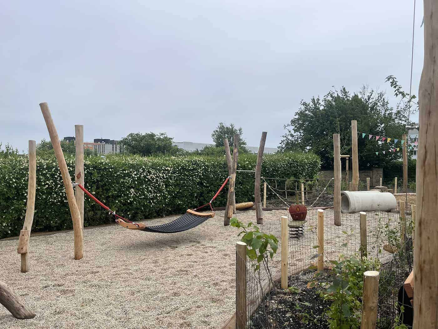 Eine große schwarze Seil-Hängematte ist zwischen robusten Holzstämmen auf einem Spielplatz mit hellem Kiesuntergrund gespannt. Im Hintergrund sind eine Betonröhre und ein rotes Erdbeer-Spielgerät zu sehen.