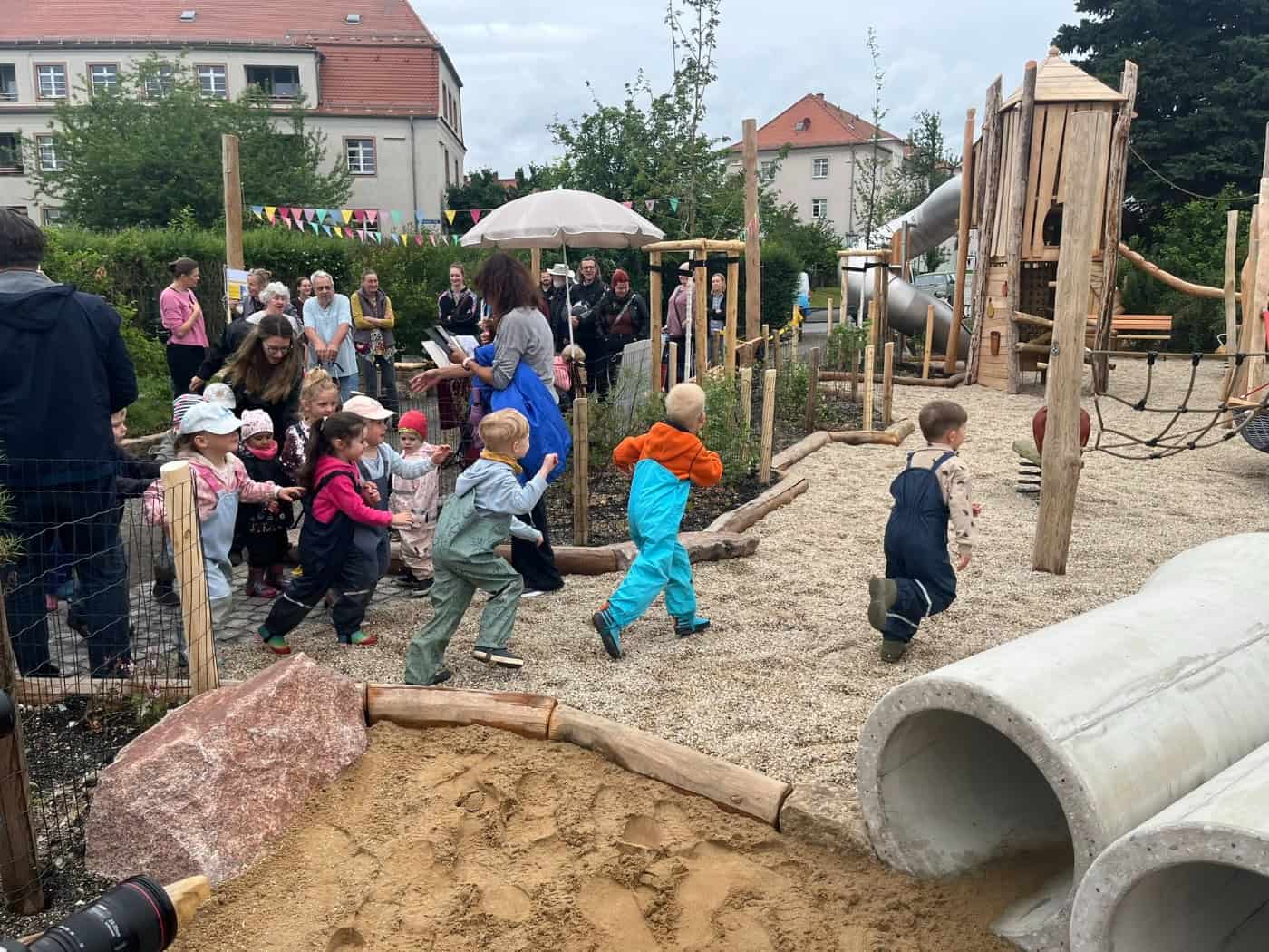 Mehrere kleine Kinder in bunter Kleidung und Matschhosen rennen voller Vorfreude über den frischen Kies des neu geöffneten Spielplatzes am Frauensteiner Platz in Dresden. Im Vordergrund sieht man eine Sandkiste und Betonröhren.