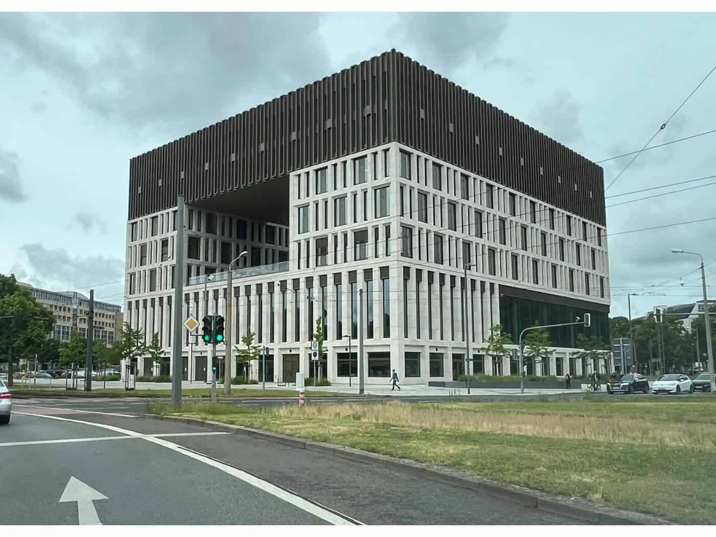 Außenansicht des neuen, modernen Stadtforums in Dresden an einer mehrspurigen Straße bei bewölktem Himmel. Das markante, kubische Verwaltungsgebäude besticht durch seine helle Steinfassade, eine große quadratische Aussparung im oberen Bereich und eine dunkle Dachkrone. Im Vordergrund ist eine Straße