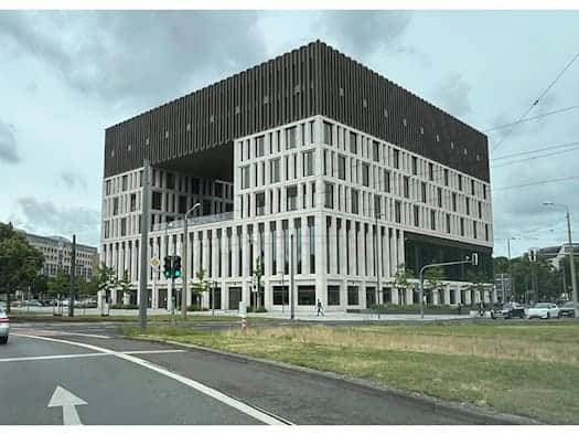Außenansicht des neuen, modernen Stadtforums in Dresden an einer mehrspurigen Straße bei bewölktem Himmel. Das markante, kubische Verwaltungsgebäude besticht durch seine helle Steinfassade, eine große quadratische Aussparung im oberen Bereich und eine dunkle Dachkrone. Im Vordergrund ist eine Straße
