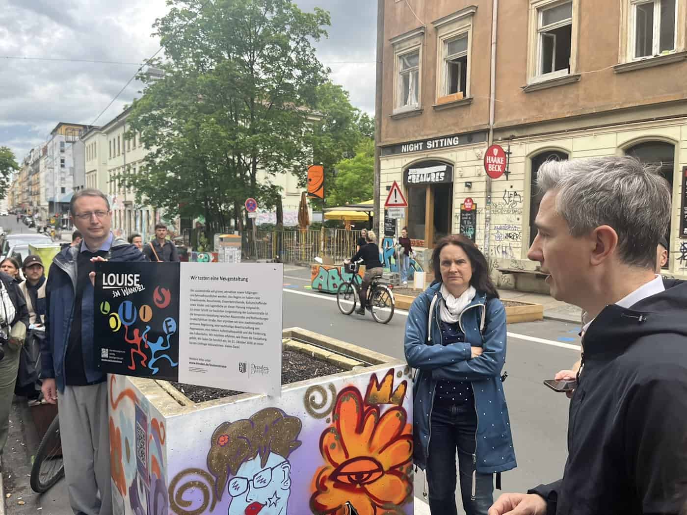 André Zschoge vom Stadtplanungsamt steht links im Bild im blauen Parka neben einem mit Graffiti verzierten Pflanzkübel und hält ein Informationsschild. Rechts im Vordergrund ist Baubürgermeister Stephan Kühn im Profil im Gespräch zu sehen.