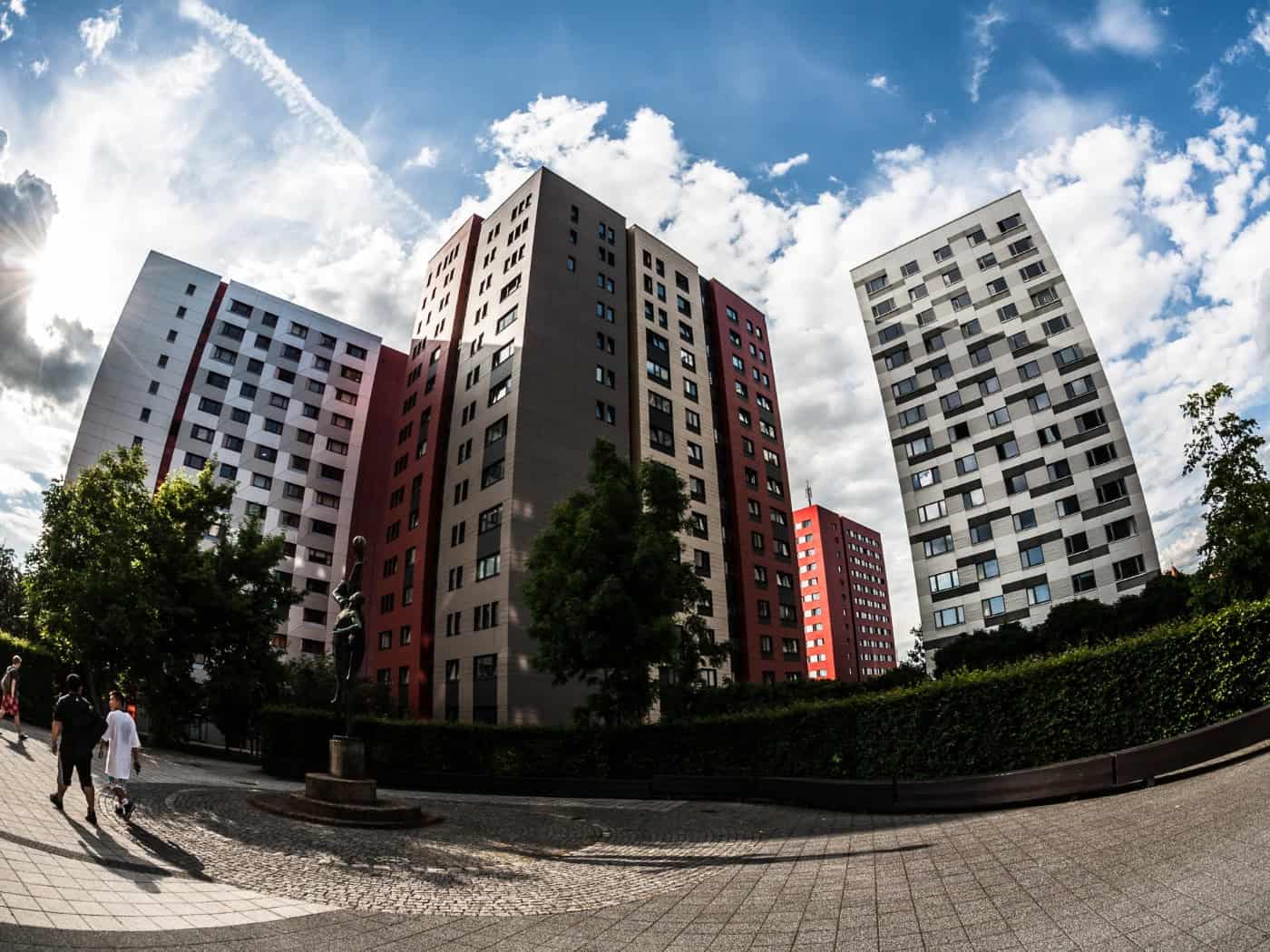 Eine Weitwinkel-Aufnahme mit Fischaugen-Effekt von mehreren modernen Hochhäusern einer Wohnheimanlage unter einem blau-weißen Wolkenhimmel. Die Fassaden sind in vertikalen Mustern aus den Farben Grau, Rot und Weiß gestaltet. 