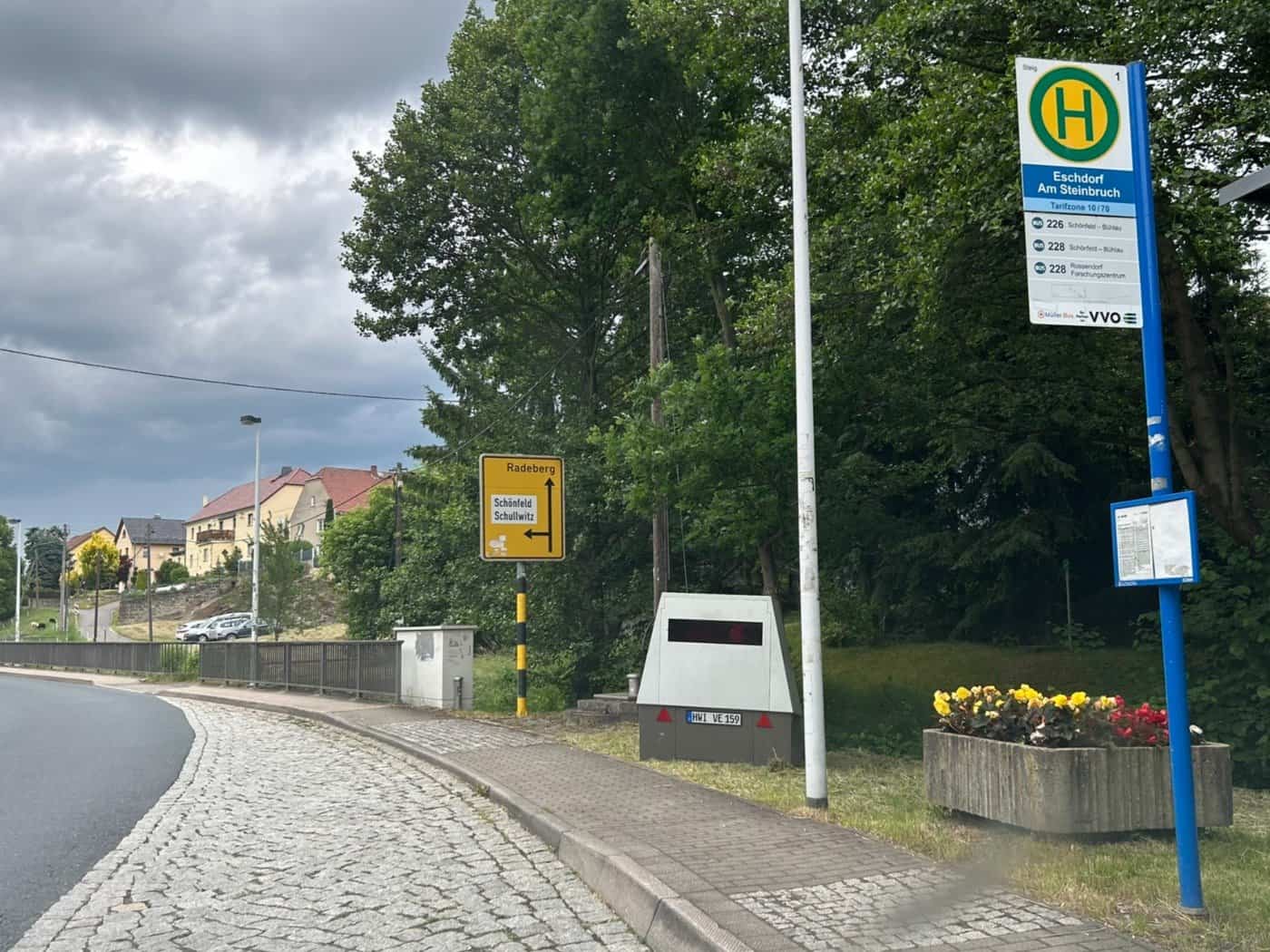 Ein grauer, mobiler Blitzer-Anhänger mit dem Kennzeichen HWI-VE 159 steht auf dem Gehweg neben dem blauen Schild der Bushaltestelle „Eschdorf Am Steinbruch“. Im Hintergrund ist ein gelbes Ortshinweisschild nach Radeberg zu sehen.