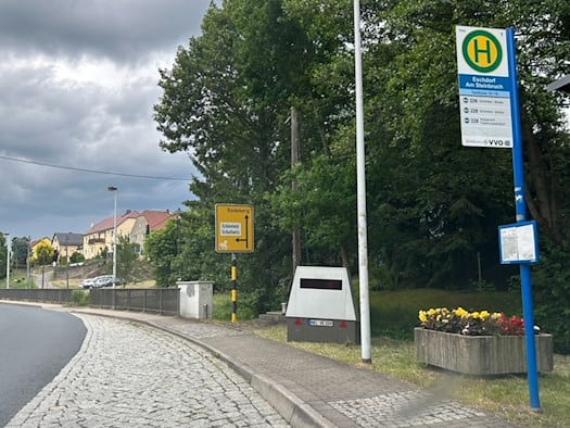 Ein grauer, mobiler Blitzer-Anhänger mit dem Kennzeichen HWI-VE 159 steht auf dem Gehweg neben dem blauen Schild der Bushaltestelle „Eschdorf Am Steinbruch“. Im Hintergrund ist ein gelbes Ortshinweisschild nach Radeberg zu sehen.
