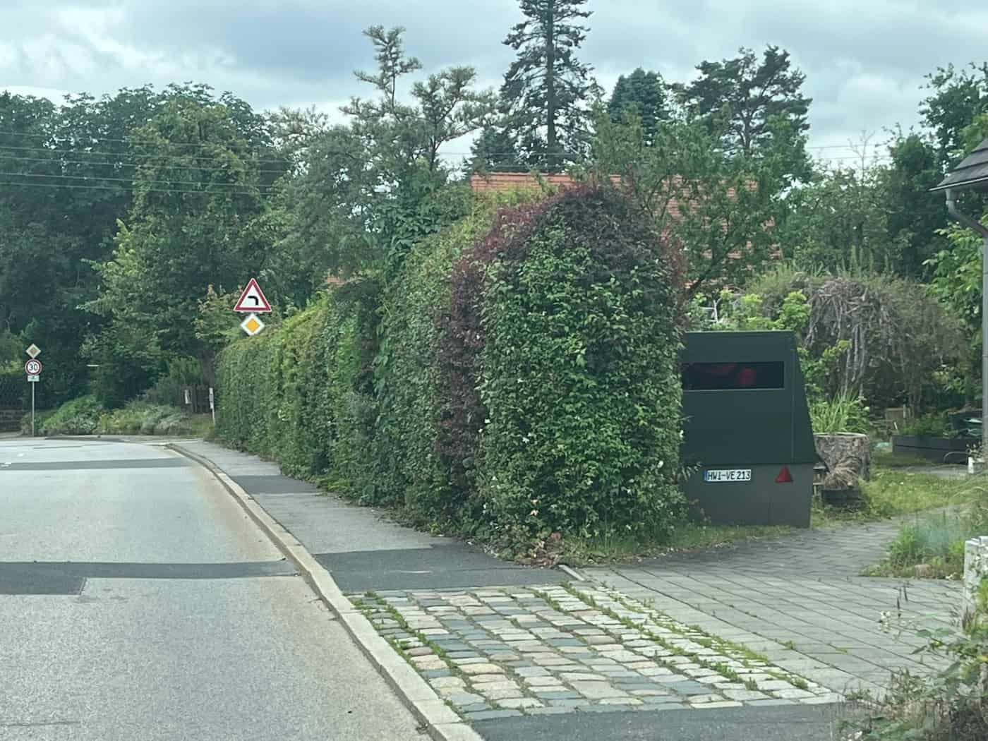 Eine Außenaufnahme eines dunkelgrünen, keilförmigen Blitzer-Anhängers (Enforcement-Trailer) mit dem Kennzeichen „HWI-VE 213“, der auf einem gepflasterten Gehwegbereich abgestellt ist. Der Blitzer steht direkt neben einer hohen, dichten grünen Hecke.