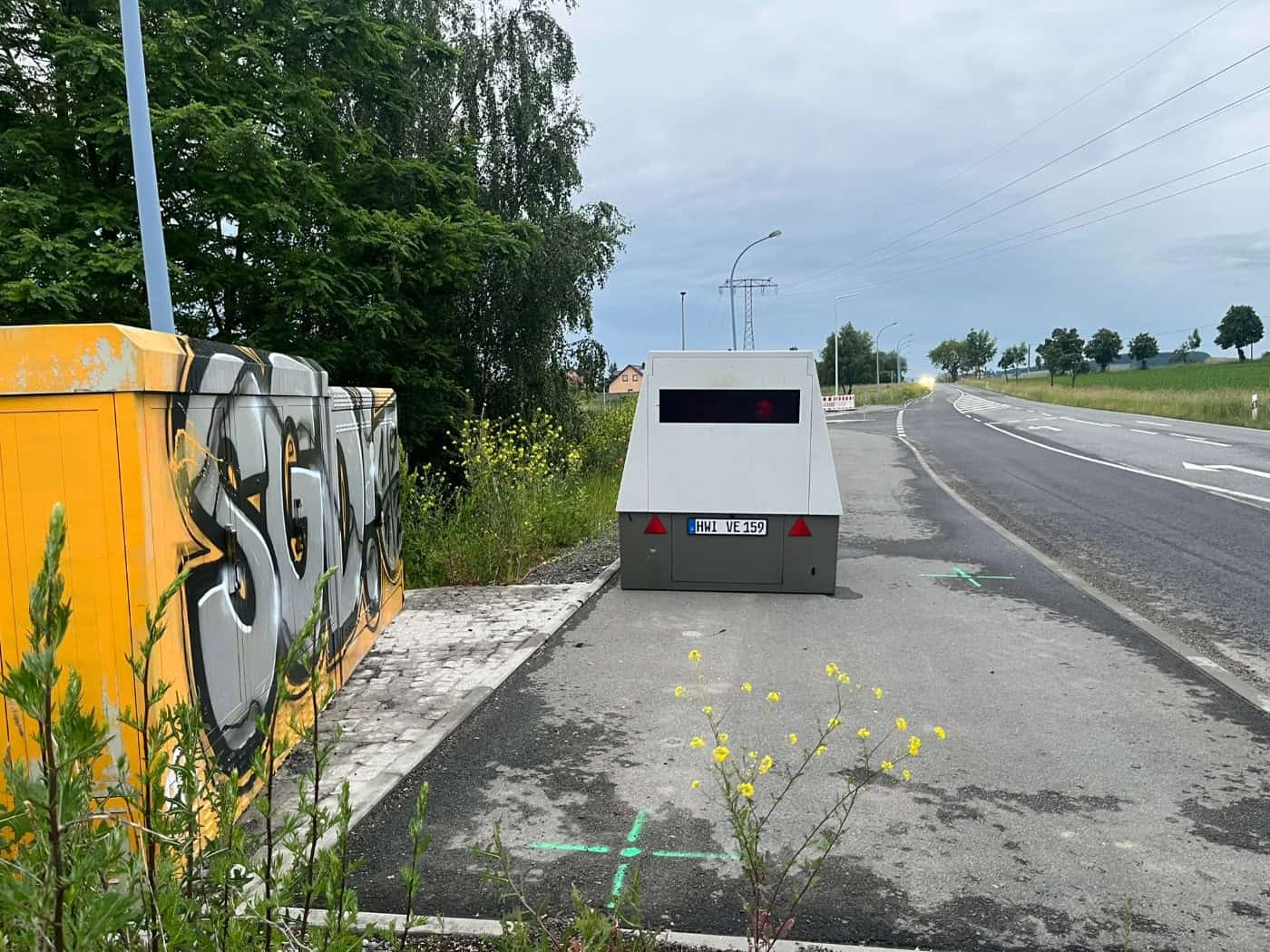 Ein grauer, mobiler Blitzer-Anhänger mit dem Kennzeichen HWI-VE 159 steht auf einer Asphaltfläche am Rand der Radeberger Landstraße. Direkt daneben befindet sich ein großer gelber Stromkasten, der mit einem auffälligen, grau-schwarzen „SGD“-Graffiti besprüht ist. Im Vordergrund blühen wilde gelbe Ra