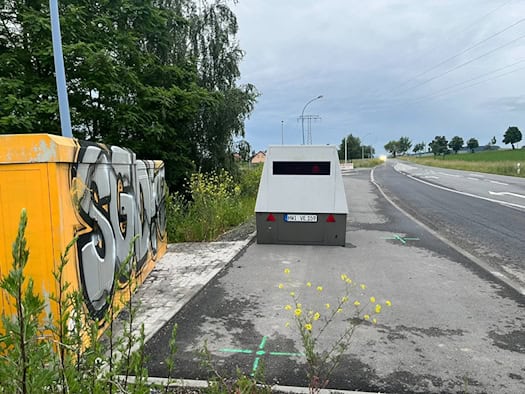 Ein grauer, mobiler Blitzer-Anhänger mit dem Kennzeichen HWI-VE 159 steht auf einer Asphaltfläche am Rand der Radeberger Landstraße. Direkt daneben befindet sich ein großer gelber Stromkasten, der mit einem auffälligen, grau-schwarzen „SGD“-Graffiti besprüht ist. Im Vordergrund blühen wilde gelbe Ra
