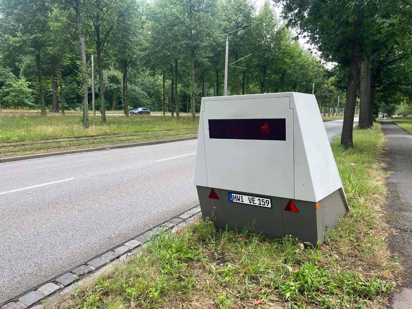 Ein grauer, anhängerbasierter Blitzer mit dem Kennzeichen HWI-VE 159 steht auf einem Grünstreifen neben einer Straße an der Stübelallee in Dresden. Im Hintergrund sind Bäume und eine Straßenbahntrasse zu sehen.