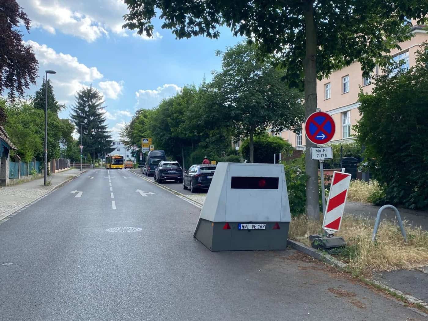 Ein grauer, kantiger Geschwindigkeitsmessanhänger mit dem Kennzeichen „HWI-VE 167“ steht am rechten Rand einer asphaltierten Wohnstraße neben einem Baum und einem Halteverbotsschild. Im Hintergrund sind Autos und ein gelber Bus zu sehen.