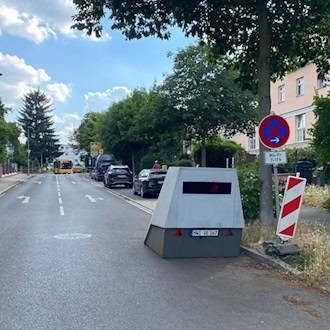 Ein grauer, kantiger Geschwindigkeitsmessanhänger mit dem Kennzeichen „HWI-VE 167“ steht am rechten Rand einer asphaltierten Wohnstraße neben einem Baum und einem Halteverbotsschild. Im Hintergrund sind Autos und ein gelber Bus zu sehen.