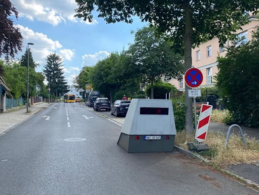 Ein grauer, kantiger Geschwindigkeitsmessanhänger mit dem Kennzeichen „HWI-VE 167“ steht am rechten Rand einer asphaltierten Wohnstraße neben einem Baum und einem Halteverbotsschild. Im Hintergrund sind Autos und ein gelber Bus zu sehen.