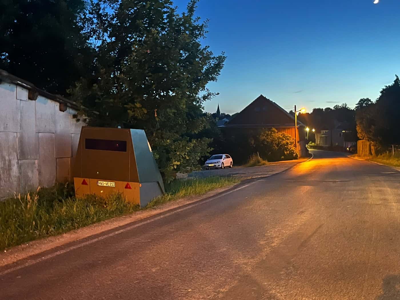 Ein olivgrüner, gepanzerter Blitzer-Anhänger mit dem Kennzeichen HWI-VE 213 steht in der Dämmerung am Grasrand einer Landstraße. Im Hintergrund beleuchtet eine Straßenlaterne ein großes ländliches Gebäude.