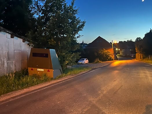 Ein olivgrüner, gepanzerter Blitzer-Anhänger mit dem Kennzeichen HWI-VE 213 steht in der Dämmerung am Grasrand einer Landstraße. Im Hintergrund beleuchtet eine Straßenlaterne ein großes ländliches Gebäude.