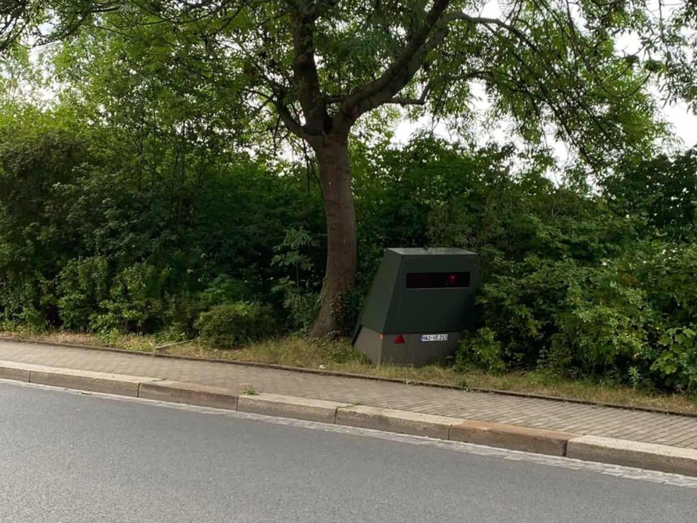 Ein olivgrüner, teilstationärer Blitzer-Anhänger steht am Fahrbahnrand unter einem grünen Laubbaum vor einer dichten Hecke.