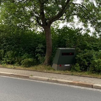 Ein olivgrüner, teilstationärer Blitzer-Anhänger steht am Fahrbahnrand unter einem grünen Laubbaum vor einer dichten Hecke.