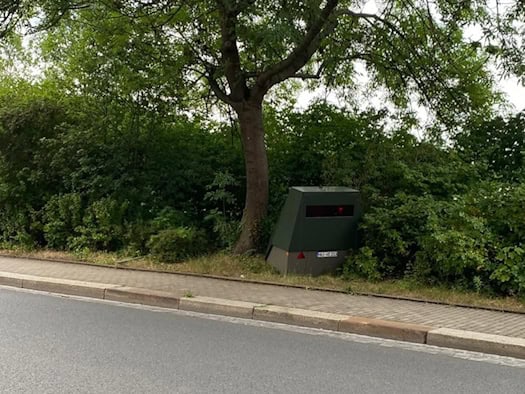 Ein olivgrüner, teilstationärer Blitzer-Anhänger steht am Fahrbahnrand unter einem grünen Laubbaum vor einer dichten Hecke.