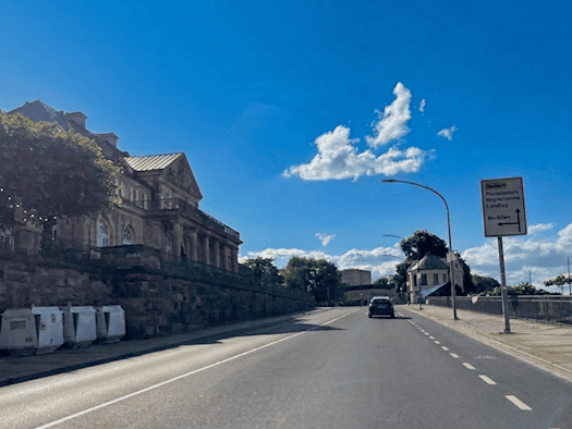 Blick auf die leere, asphaltierte Straße des Dresdner Terrassenufers mit der Sandsteinmauer und dem historischen Gebäude des Italienischen Dörfchens auf der linken Seite. Ein einzelnes Auto fährt auf der Fahrbahn unter strahlend blauem Himmel, rechts steht ein gelb-weißes Wegweiserschild.