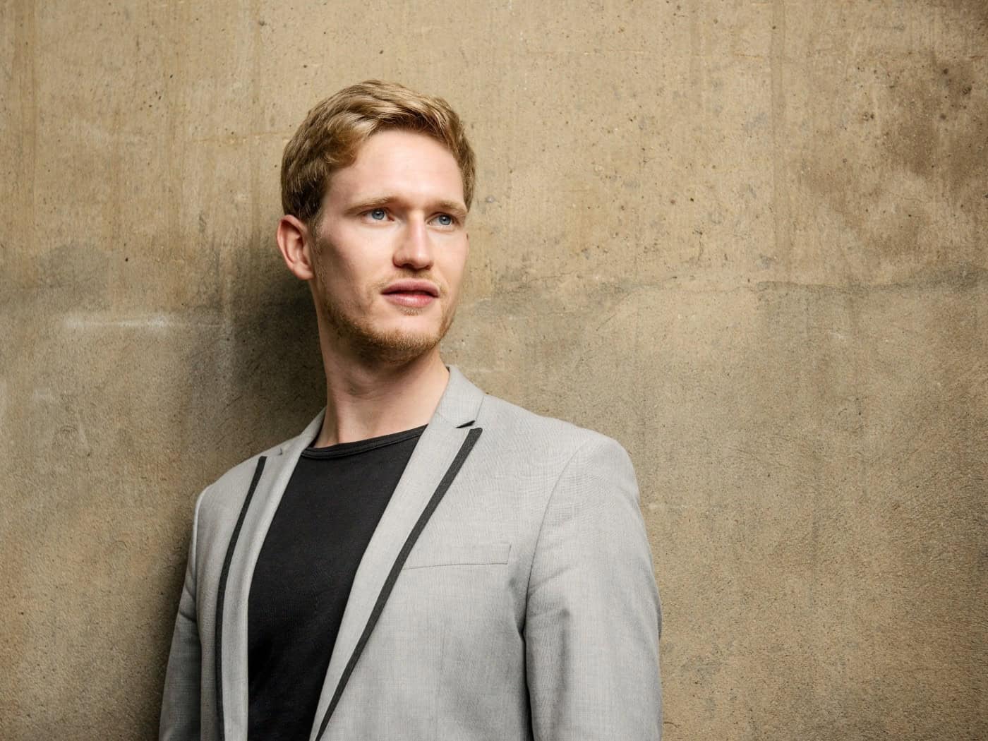 Ein professionelles Porträt des Dirigenten Thomas Guggeis in eleganter Kleidung vor einem schlichten, dezent ausgeleuchteten Hintergrund. Er blickt mit aufmerksamem, ruhigem Ausdruck direkt in die Kamera.