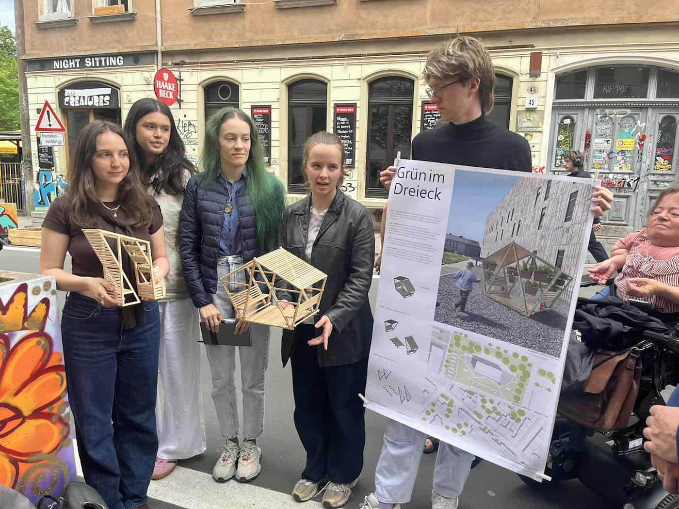 Eine Gruppe von fünf jungen Studierenden steht auf der Louisenstraße und hält zwei filigrane Modelle aus Holzstäben sowie ein großes Plakat mit der Aufschrift „Grün im Dreieck“ in die Kamera, das Entwürfe für neue Stadtmöbel zeigt.