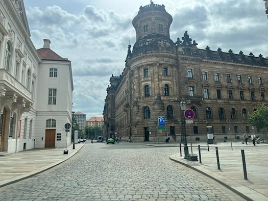 Blick über eine gepflasterte Straße auf das historische, sandsteinfarbene Gebäude der Polizeidirektion Dresden mit einem markanten runden Eckturm. Links steht ein weißes, klassizistisches Gebäude unter bewölktem Himmel.
