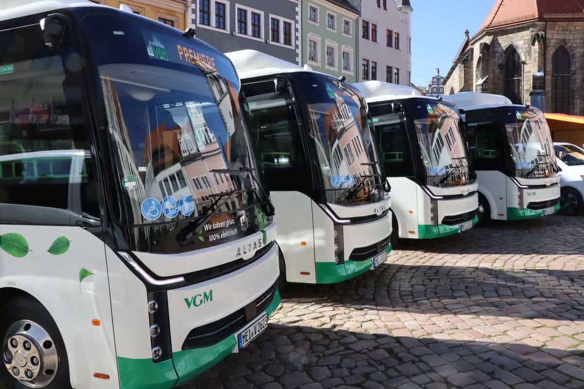 Foto: Vier grün-weiße Personenbusse stehen nebeneinander auf einem Platz mit historischen Gebäuden