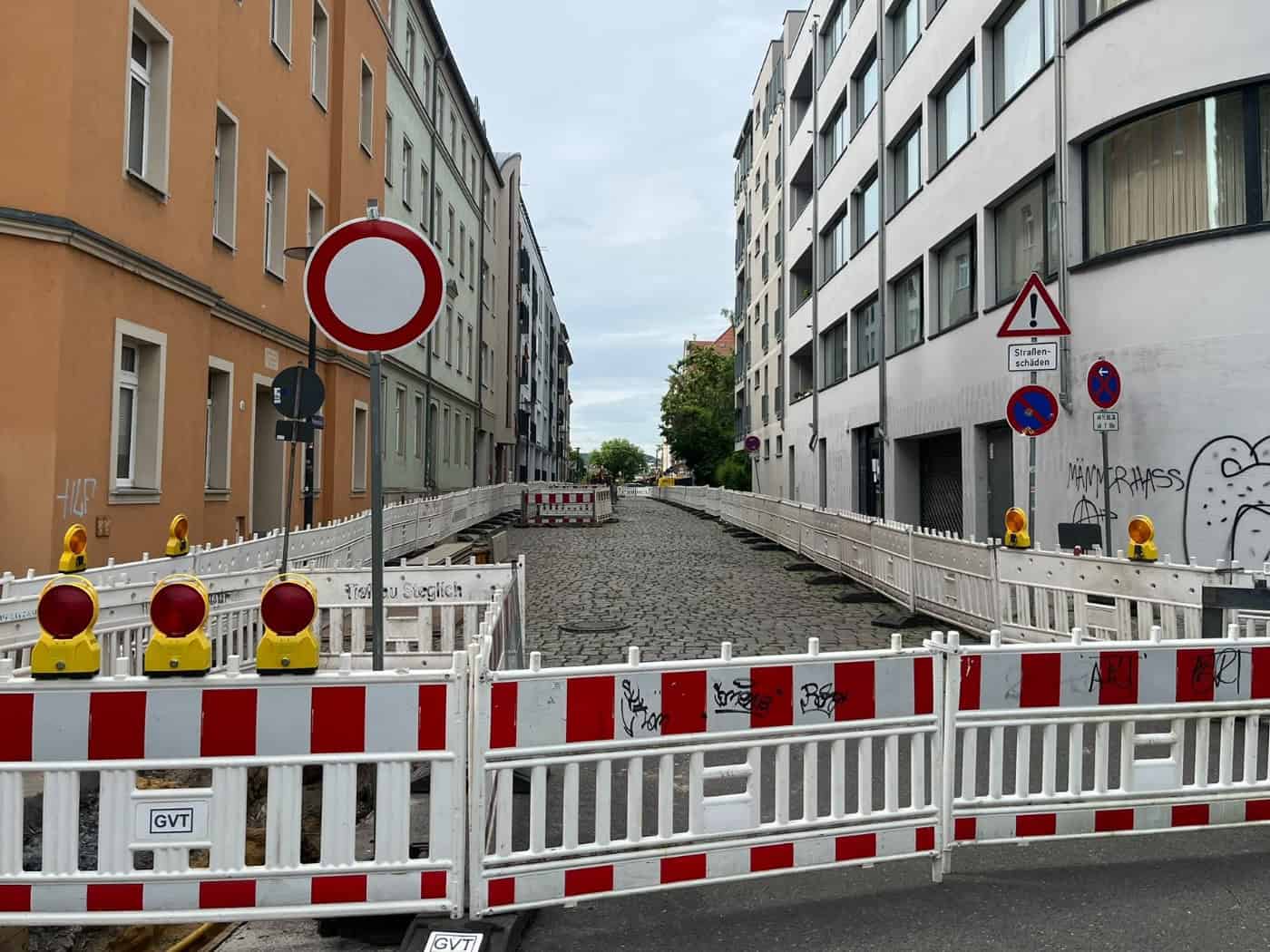 lick entlang der von mehrgeschossigen Wohnhäusern gesäumten Vorwerkstraße in Dresden-Friedrichstadt.  Die Fahrbahn besteht aus altem, unebenem Natursteinpflaster und ist mit weißen Baustellengittern abgesperrt.  Im Vordergrund steht eine rot-weiße Absperrschranke mit gelben Warnleuchten.