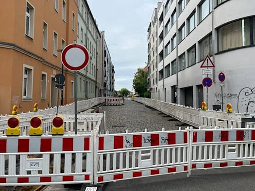 lick entlang der von mehrgeschossigen Wohnhäusern gesäumten Vorwerkstraße in Dresden-Friedrichstadt.  Die Fahrbahn besteht aus altem, unebenem Natursteinpflaster und ist mit weißen Baustellengittern abgesperrt.  Im Vordergrund steht eine rot-weiße Absperrschranke mit gelben Warnleuchten.