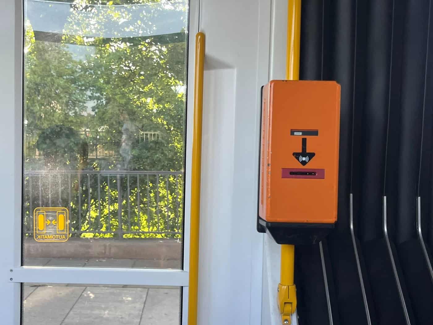 Ein orangefarbener, kastenförmiger Fahrkartenentwerter ist an einer gelben Haltestange im Innenraum einer Straßenbahn montiert. Links daneben befindet sich ein großes Fenster mit Blick auf grüne Bäume.