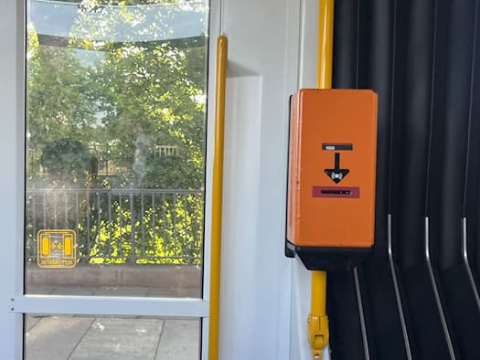 Ein orangefarbener, kastenförmiger Fahrkartenentwerter ist an einer gelben Haltestange im Innenraum einer Straßenbahn montiert. Links daneben befindet sich ein großes Fenster mit Blick auf grüne Bäume.