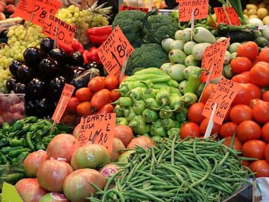 Ein reich bestückter Marktstand voller frischem Gemüse. Zu sehen sind leuchtend rote Tomaten, grüne Bohnen, Brokkoli, Paprika und dunkle Auberginen. In den Auslagen stecken orangefarbene Preisschilder mit handschriftlichen Notizen.