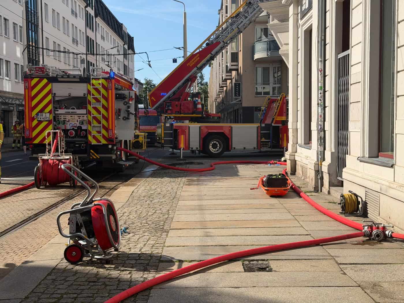 Ein rotes Löschfahrzeug und ein Drehleiterwagen der Feuerwehr Dresden stehen auf den Schienen einer gesperrten Straße. Auf dem Gehweg im Vordergrund liegen ausgerollte rote Feuerwehrschläuche, ein roter Druckbelüfter sowie eine orangefarbene Rettungstrage vor einer Häuserwand.