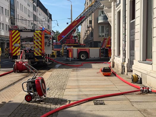 Ein rotes Löschfahrzeug und ein Drehleiterwagen der Feuerwehr Dresden stehen auf den Schienen einer gesperrten Straße. Auf dem Gehweg im Vordergrund liegen ausgerollte rote Feuerwehrschläuche, ein roter Druckbelüfter sowie eine orangefarbene Rettungstrage vor einer Häuserwand.