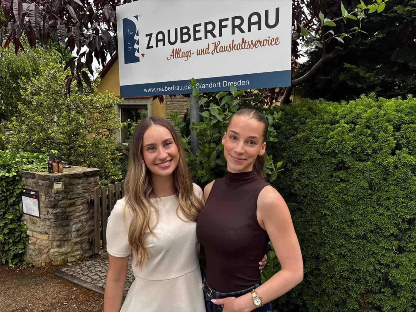 Porträt von zwei lächelnden jungen Frauen, Talia Petermann und Lisa Thauer, die nebeneinander im Freien vor einem grünen Hintergrund aus Sträuchern stehen. Sie tragen helle, sommerliche Alltagskleidung und blicken freundlich direkt in die Kamera.