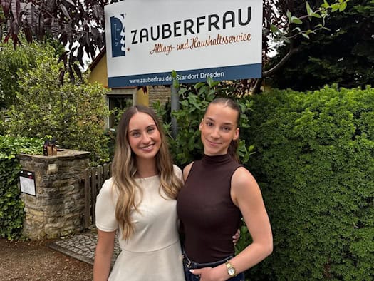 Porträt von zwei lächelnden jungen Frauen, Talia Petermann und Lisa Thauer, die nebeneinander im Freien vor einem grünen Hintergrund aus Sträuchern stehen. Sie tragen helle, sommerliche Alltagskleidung und blicken freundlich direkt in die Kamera.