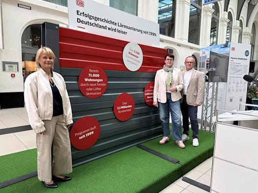 Drei Personen stehen in einer hellen Bahnhofshalle auf Kunstrasen vor einem rot-grauen Lärmschutzelement mit roten Info-Kreisen. Links eine Frau in heller Jacke, rechts eine Frau mit Brücke/Schal und ein Mann im hellen Sakko. Ein Schild zeigt das DB-Logo.