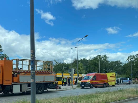 Blick aus einem Fahrzeug auf eine Unfallstelle in Dresden: Ein orangefarbenes Rettungsfahrzeug, ein roter Einsatzleitwagen und die Polizei stehen an einer gelben Straßenbahn. Rechts unten spiegelt sich die Straße im Außenspiegel.